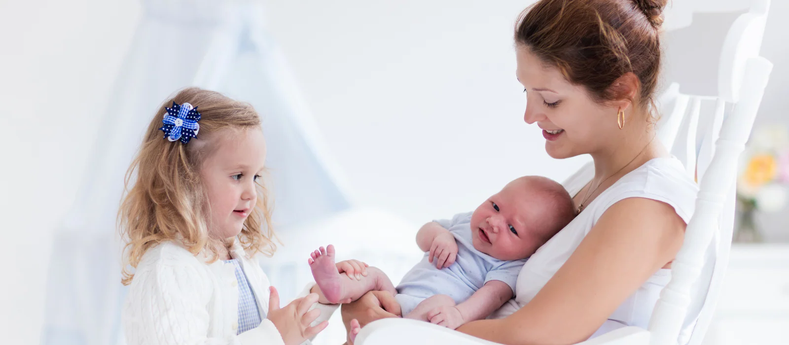 Mother holding a newborn while a toddler stands nearby during a busy day at home